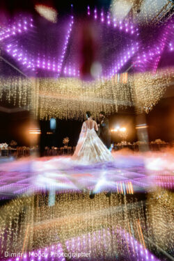 A bride and groom share their first dance under purple and gold lights in a decorated wedding venue.