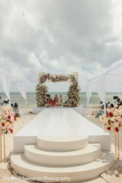 A decorated wedding altar and white aisle set up on a beach with people seated on either side under canopies.