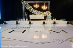 White stage with a white couch, floral arch, chandeliers, and "H & M" monogram on the floor.