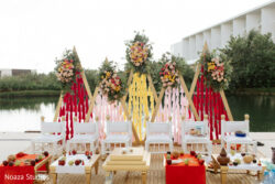 A decorative outdoor setup with white chairs, floral arrangements, and ritual items near a pond.