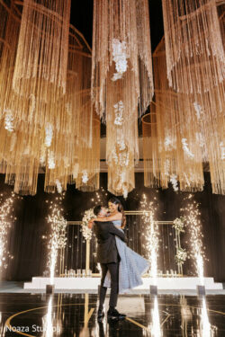A couple embraces on a black stage with golden hanging decorations and sparklers surrounding them.