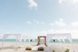 White canopies and floral arch set up for a wedding ceremony on a sandy beach with the ocean in the background.