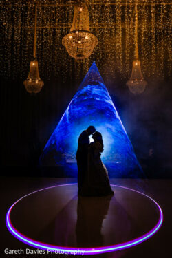 A couple stands in silhouette embracing on a lit dance floor with chandeliers and a blue triangular light backdrop.