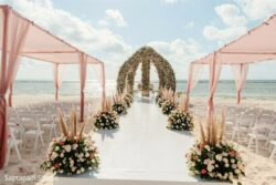 A wedding altar with floral arches and pink drapes is set up on a beach with rows of white chairs and flower arrangements.