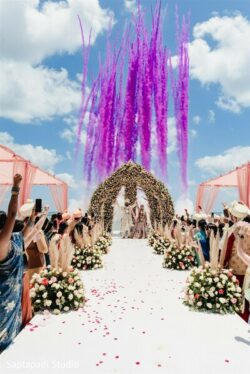 A wedding couple stands under a floral arch as purple smoke rises in the sky; guests are cheering along a flower-lined aisle.