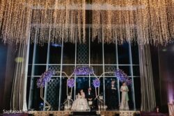 A wedding ceremony with a couple seated on stage under floral arches, flanked by two people, beneath hanging string lights.