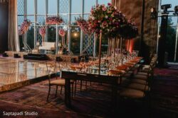 Elegant event setup with a decorated stage, floral arrangements, and a long dining table with glassware and plates.