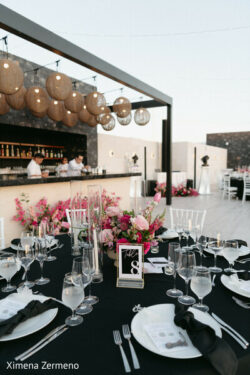 A formal outdoor event table set with glassware and black napkins, with a bar and string lights in the background.