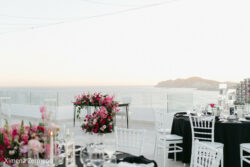 Outdoor event setup with white chairs, black tablecloths, and pink floral arrangements overlooking the ocean at sunset.