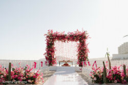 Outdoor wedding altar with a pink and red floral arch, white chairs, and aisle decorations under a clear sky.