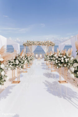 Elegant beach wedding setup with white floral arrangements, a glossy aisle, and an ocean backdrop under a clear blue sky.