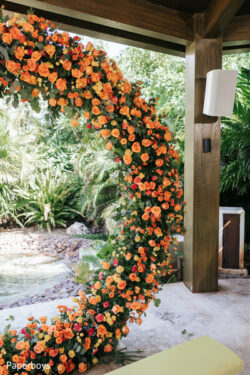 A large crescent-shaped floral arrangement with orange roses stands outdoors near a wooden structure and lush greenery.