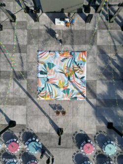 default Aerial view of a colorful canopy in a plaza, surrounded by round tables with chairs and decorative string lights.