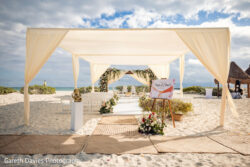 Outdoor beach wedding setup with a white canopy, chairs, floral arrangements, and a sign reading "Pepi & Chris.
