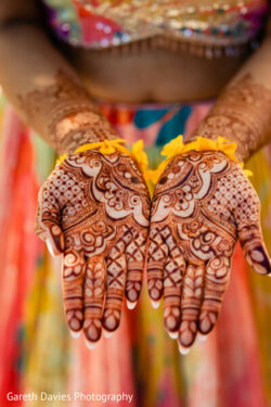 Close-up of two open hands decorated with intricate henna designs, with blurred colorful clothing in the background.