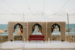 A red tufted sofa sits in front of ornate arches with hanging lanterns, set by the sea under string lights.