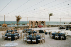 Outdoor beachfront event setup with round tables, navy linens, gold chairs, string lights, and an ornate stage backdrop.