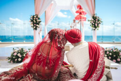 A couple in traditional Indian wedding attire sits under a canopy, facing the ocean at an outdoor ceremony.
