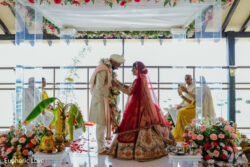 A bride and groom in traditional attire exchange garlands at a floral-decorated indoor wedding ceremony.