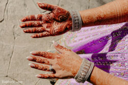 Two hands with intricate henna designs, silver bangles, and a ring, posed above a lilac and silver patterned dress.
