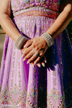 A woman in a detailed pink and purple dress stands with her hands clasped, displaying henna and bangles.