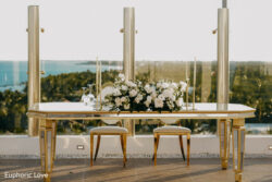 A gold table with two chairs, white floral centerpiece, and candlesticks set on a terrace overlooking greenery and water.